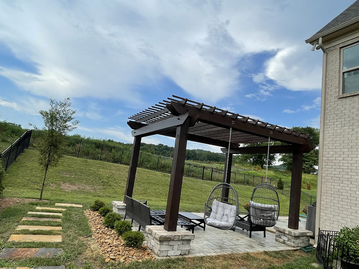 A dark-stained wood pergola over a paver patio with a hillside view