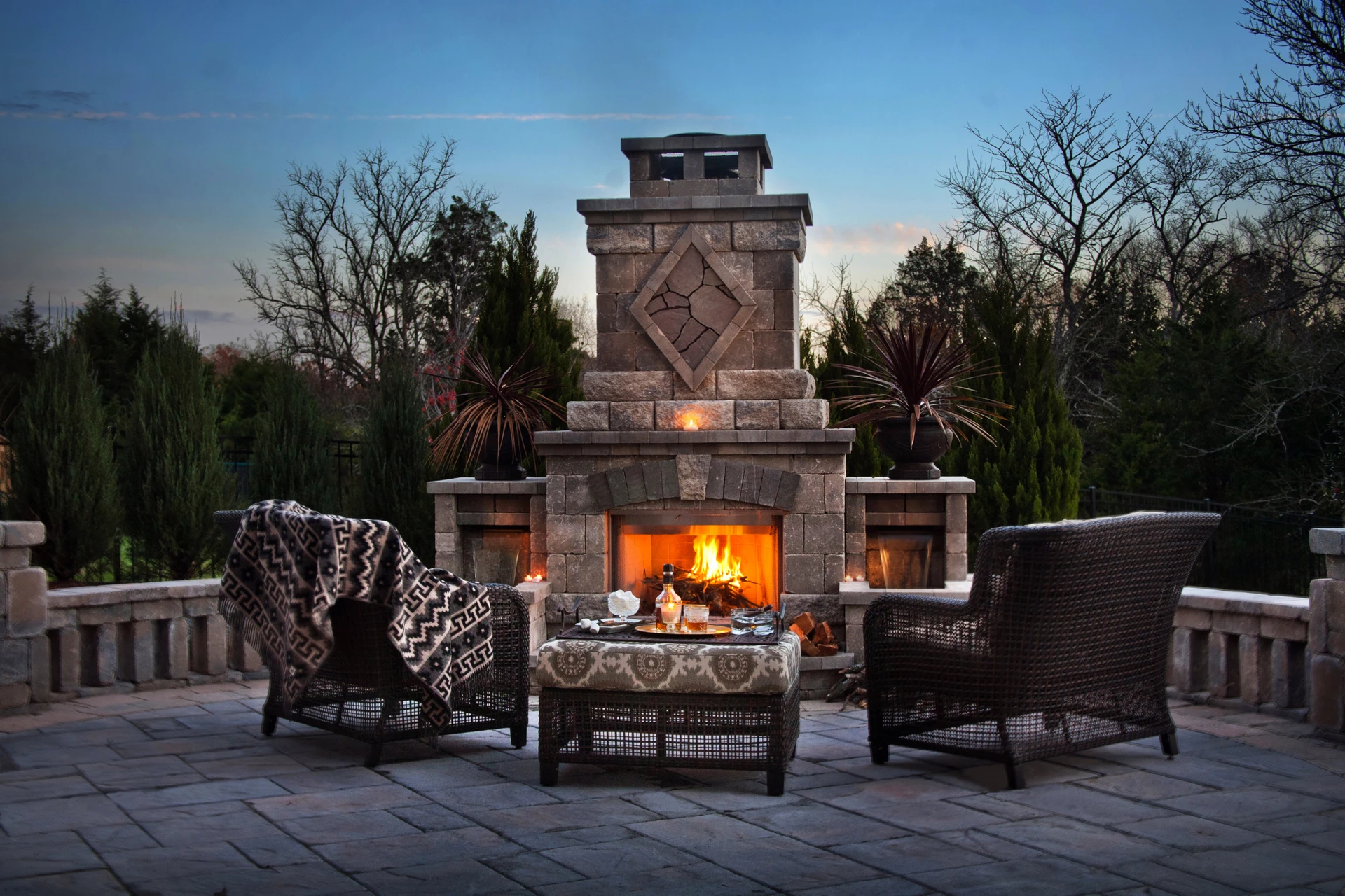 A grand stone outdoor fireplace and lounge on a flagstone terrace at dusk